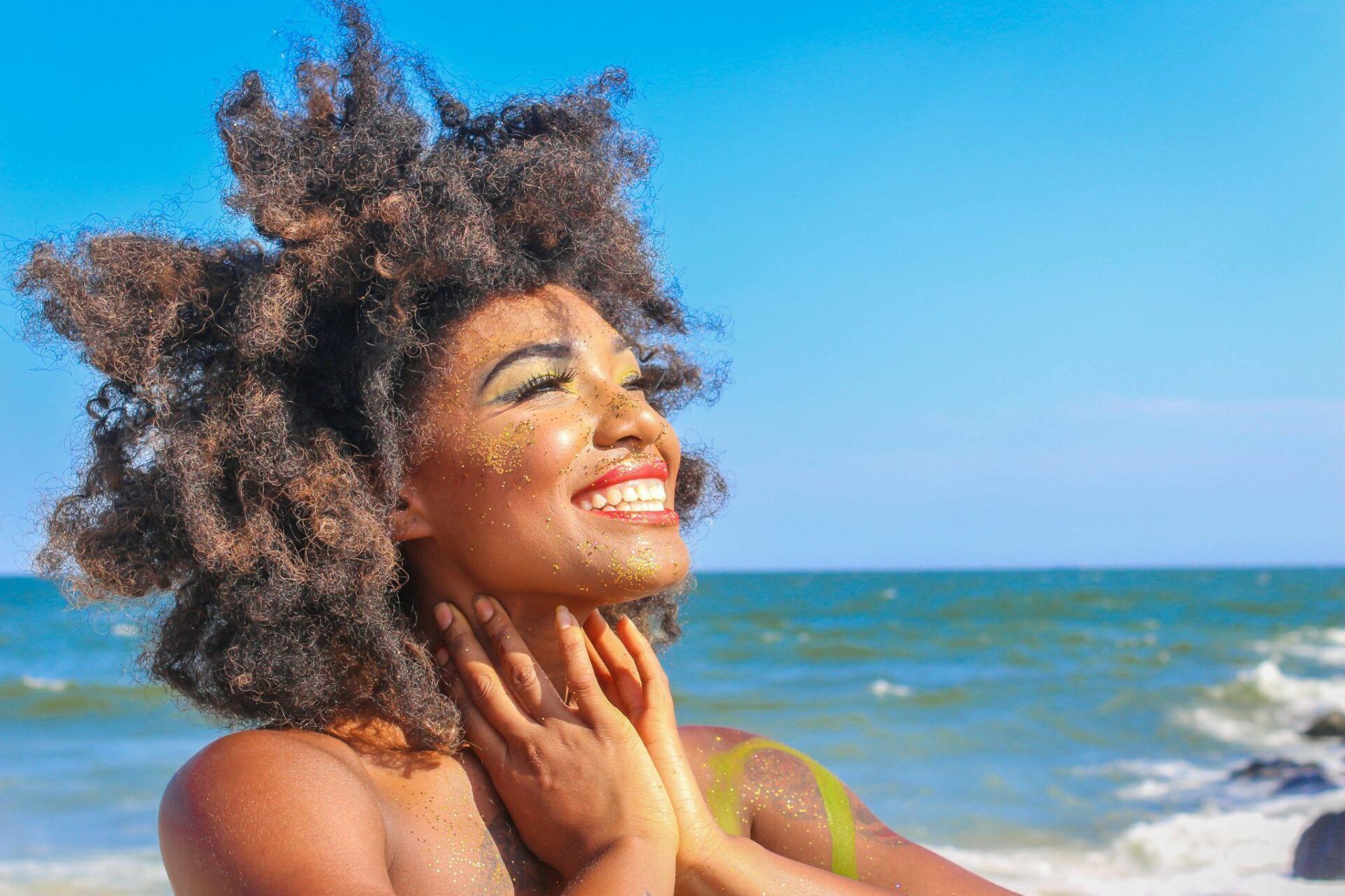 A close-up photo of a woman with glittering make-up and waxed upper lip. She basks in the sun on the beach.